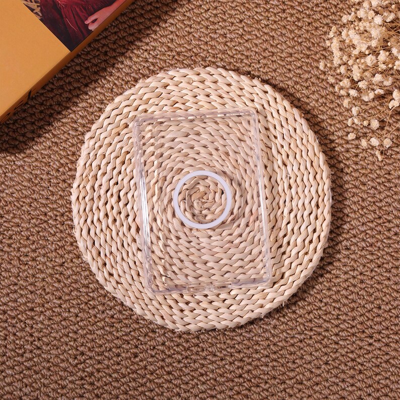 May include: A clear, rectangular phone case with a circular, white ring in the center. The case is placed on a round, woven straw mat, set against a brown, textured background. The overall aesthetic is natural and minimalist.