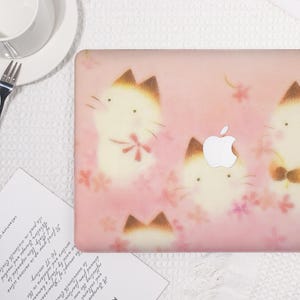 K&ouml;nnte beinhalten: Eine rosa Laptop-H&uuml;lle mit einem sich wiederholenden Muster aus Cartoon-Katzen mit braunen Ohren und rosa Blumendetails. Das Apple-Logo ist sichtbar. Die H&uuml;lle liegt auf einer wei&szlig;en Oberfl&auml;che.