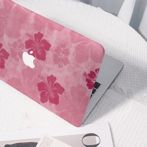 May include: A pink laptop case featuring a repeating hibiscus flower pattern in varying shades of pink. The Apple logo is visible. The case is on a white surface.