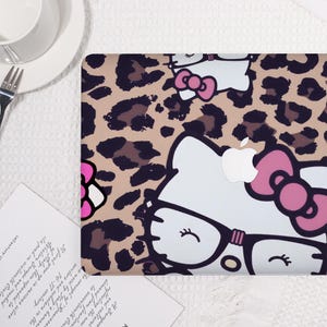 Pu&ograve; includere: Una custodia per laptop con motivo leopardato e design Hello Kitty. La custodia ha uno sfondo beige con macchie marroni e un volto di Hello Kitty bianco con dettagli rosa e occhiali.