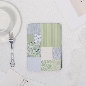May include: A rectangular coaster with a patchwork design. The coaster features various patterns in shades of green and blue, including stripes, floral prints, and geometric shapes. A white plate, bowl, and fork are in the background.