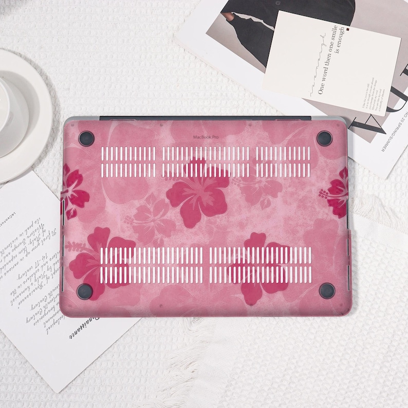 May include: A pink laptop case with a floral hibiscus pattern. The case has a textured background and a series of ventilation slots. The design features large, dark pink hibiscus flowers. The case is designed for a MacBook Pro.