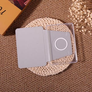 May include: A gray tablet case with a clear plastic frame. The case is open, revealing a light gray interior with a circular cutout. The case is resting on a woven mat, with a book and dried flowers in the background.