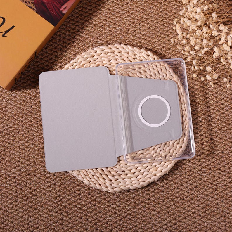 May include: A gray tablet case with a clear plastic frame. The case is open, revealing a light gray interior with a circular cutout. The case is resting on a woven mat, with a book and dried flowers in the background.