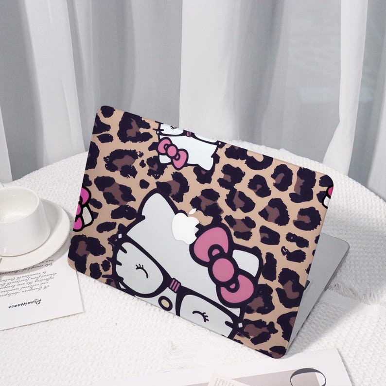 Pu&ograve; includere: Custodia per laptop con motivo leopardato e un gatto dei cartoni animati con occhiali e fiocco rosa. La custodia &egrave; beige, marrone, bianca e rosa. Il laptop &egrave; su una superficie bianca.