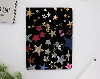 Può includere: Una copertina per quaderno nera con un motivo a stelle colorate. Le stelle sono di diverse dimensioni e colori, tra cui rosso, blu, oro e argento.