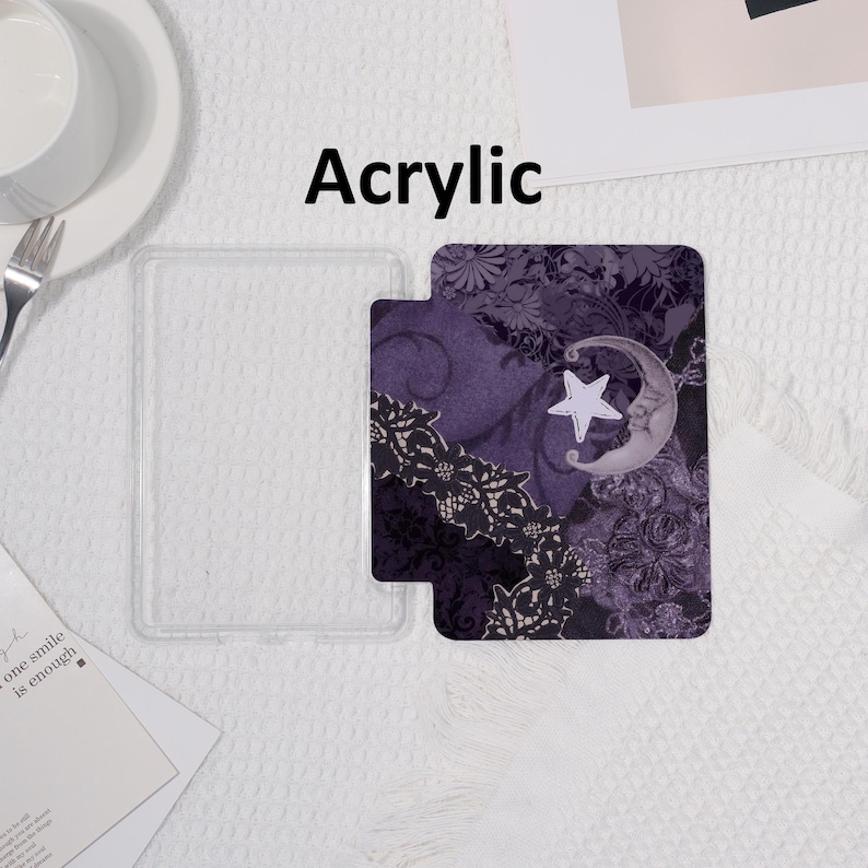 K&ouml;nnte beinhalten: Transparente Handyh&uuml;lle aus Acryl mit dekorativem Einsatz. Der Einsatz zeigt ein lila Blumenmuster mit Mond und Stern. Das Wort "Acrylic" ist in Schwarz gedruckt.