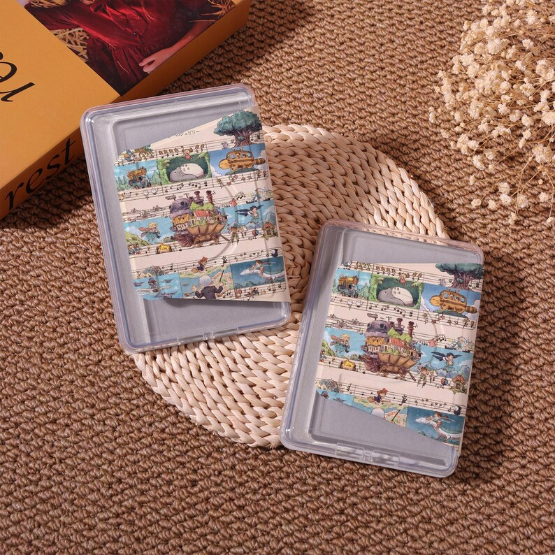 May include: Two clear plastic cases with decorative tape featuring a whimsical illustration of a fantasy landscape with musical notes. The cases are rectangular and sit on a woven mat.