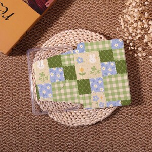 May include: A clear phone case with a colorful patchwork design. The design features squares of green gingham, blue floral patterns, and beige squares with bunny and flower illustrations. The case is resting on a woven mat.