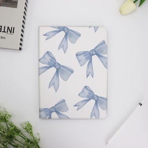 Pode incluir: Uma capa de caderno branca com um padrão de laços em aquarela azul claro. Os laços são uniformemente espaçados na capa. Uma caneta stylus branca e um livro também são visíveis.