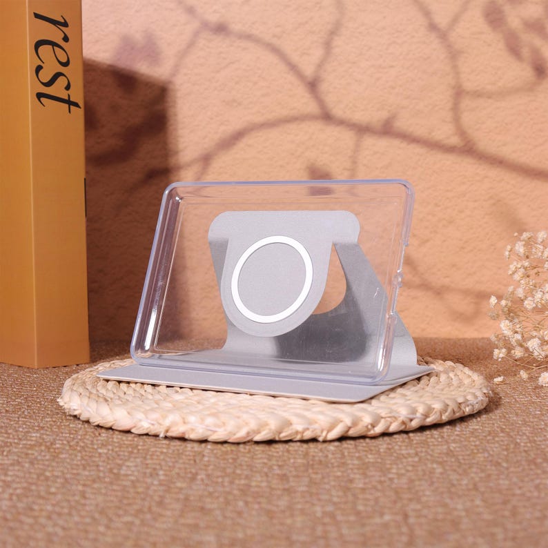May include: A clear plastic and gray tablet stand with a circular design in the center. The stand is on a woven mat, with a book that says "rest" in the background.