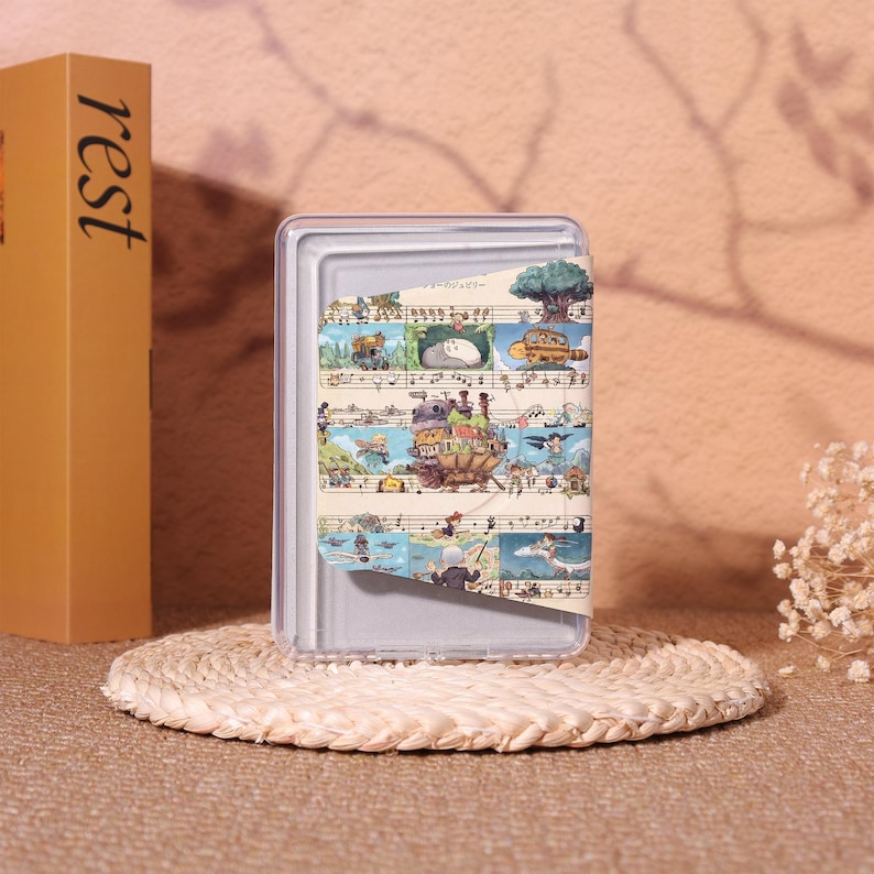 May include: A clear, rectangular phone case with a colorful illustration of a fantasy scene. The case is resting on a woven, circular coaster. A tan book with the word "rest" is visible.