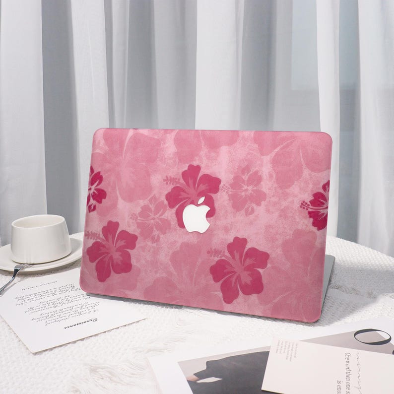 May include: Pink laptop case with a floral hibiscus pattern. The case features a white Apple logo and a repeating design of pink hibiscus flowers on a light pink background. The laptop is on a white table.