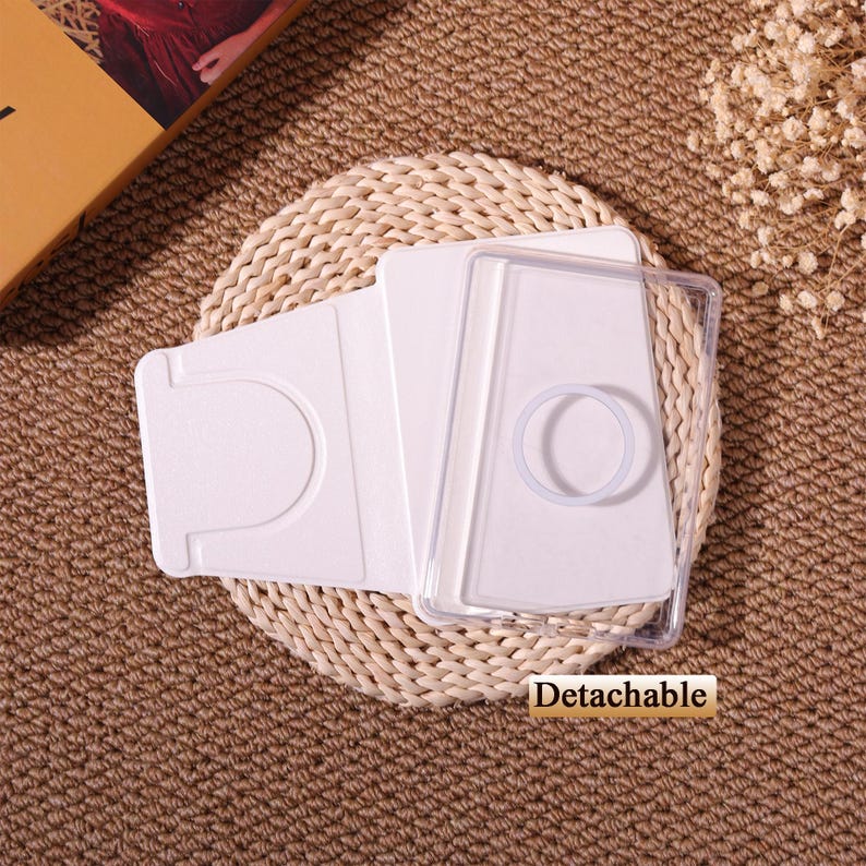 May include: A white phone case with a clear, detachable cover. The case has a curved design with a circular ring and a slot for cards. The word "Detachable" is printed on the bottom right corner of the image. The case is on a woven mat.