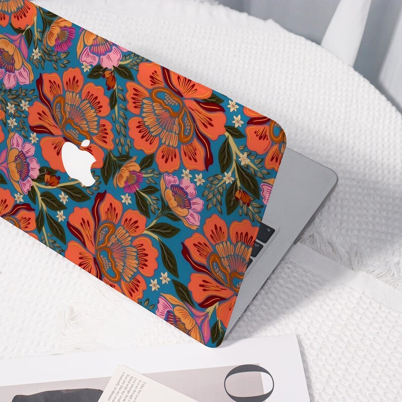 K&ouml;nnte beinhalten: Ein Laptop mit einer H&uuml;lle mit floralem Design. Die H&uuml;lle zeigt gro&szlig;e orangefarbene und rosa Bl&uuml;ten mit gr&uuml;nen Bl&auml;ttern auf einem tealfarbenen Hintergrund. Das Apple-Logo ist sichtbar. Der Laptop befindet sich auf einer wei&szlig;en Oberfl&auml;che.