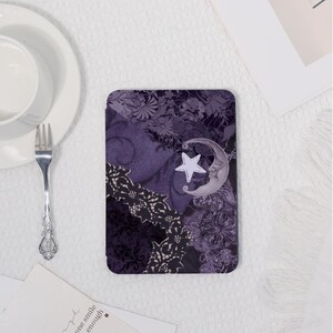K&ouml;nnte beinhalten: Ein rechteckiger Untersetzer mit einem lila-schwarzen Blumenmuster, das einen Mond und einen Stern zeigt. Der Untersetzer hat ein dekoratives Spitzendesign und einen wei&szlig;en Stern. Der Hintergrund hat ein Blumenmuster.