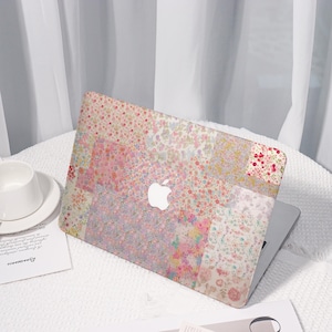 Op de afbeelding: Een laptop met een bloemenpatroon hoesje in roze, witte en gele tinten. De hoes heeft een patchwork ontwerp met een wit Apple logo. De laptop staat open op een wit oppervlak.