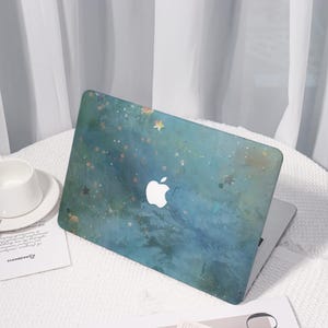 Könnte beinhalten: Ein Laptop mit einer teal- und blau-galaktischen Hülle mit einem weißen Apple-Logo. Das Design umfasst Sterne und einen Aquarelleffekt. Der Laptop befindet sich auf einer weißen Oberfläche, neben einer weißen Tasse und Untertasse.