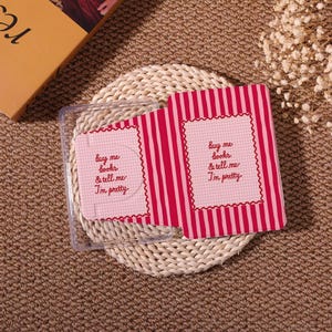 May include: A pink and white striped notebook with the text "Buy me books & tell me I'm pretty" on a pink gingham background. The notebook is open, revealing the inside pages. The notebook is on a woven mat.