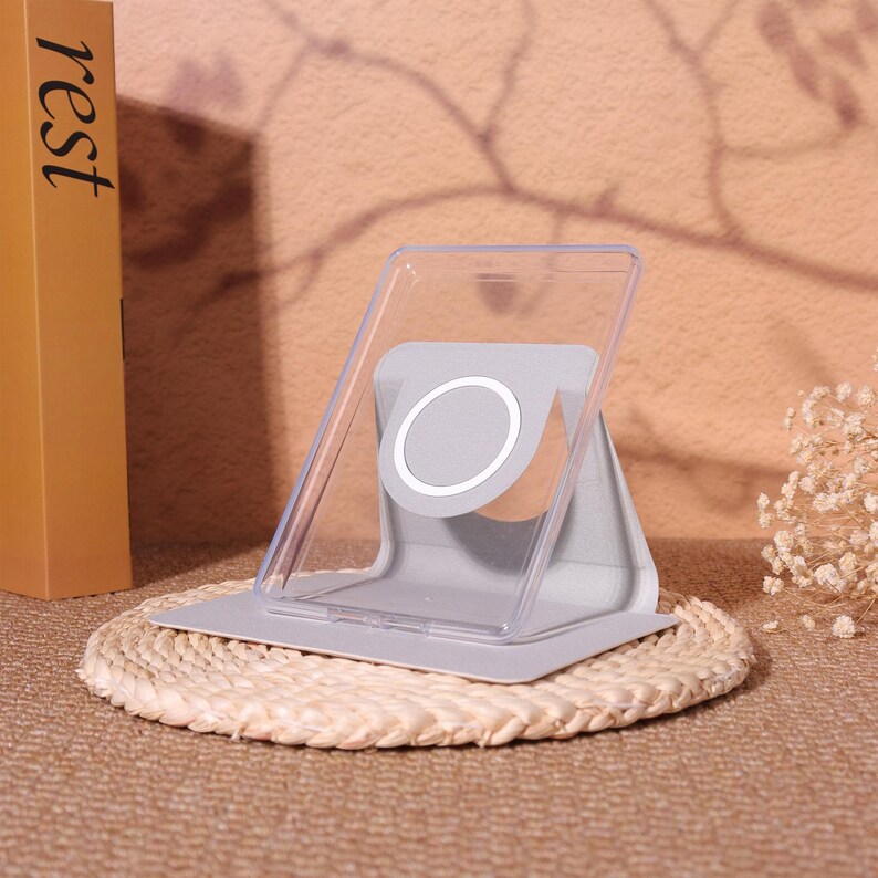 May include: A gray and clear phone stand with a white circular design. The stand is on a woven mat. A book with the word "rest" is visible in the background.