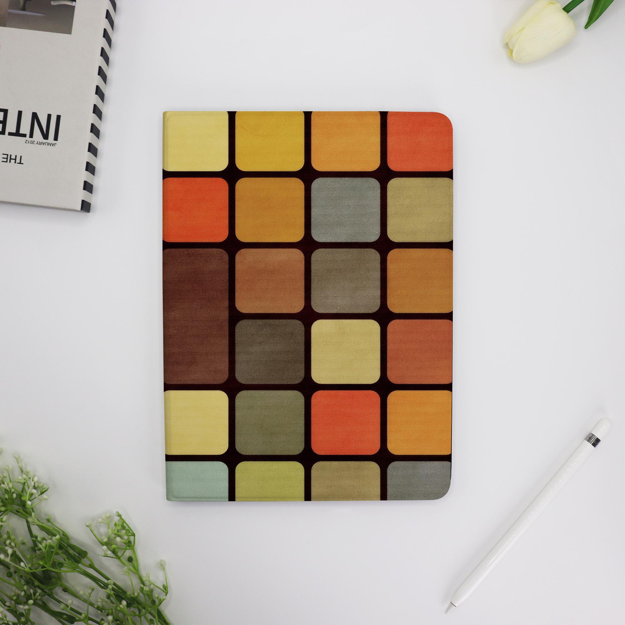 May include: A notebook with a colorful, geometric pattern of squares in shades of yellow, orange, brown, and green. The cover has a dark brown border. A white pen and a book are on the white surface.