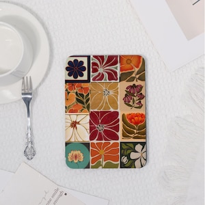 May include: A square coaster with a floral design. The coaster is divided into twelve squares, each featuring a different flower in shades of orange, red, yellow, and white. A blue flower is in the top left corner. The coaster is on a white surface.