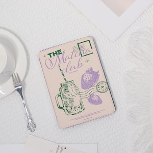 May include: A rectangular, light-colored case featuring a green and purple illustration of a mason jar with a straw, strawberries, and the text "The Matcha Club." The case has a postage stamp design and the words "Est. 2016."