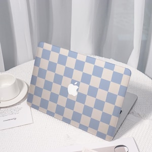 Pode incluir: Um laptop com uma capa com padrão xadrez azul claro e bege. O logotipo da Apple é visível no centro. O laptop está aberto, revelando a tela. À esquerda, há uma xícara e um pires brancos.