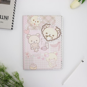 Könnte beinhalten: Ein rosa-beiges Notizbuch mit Cartoon-Bären-Illustrationen. Das Cover hat ein Patchwork-Design mit Herzen, Blumen und dem Text "おいでおいで". Das Notizbuch ist von einem weißen Hintergrund mit einem Stift und einer Blume umgeben.