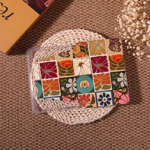 May include: A clear protective case with a colorful floral pattern. The pattern features squares with flowers in shades of red, orange, white, and blue. The case is on a woven mat.