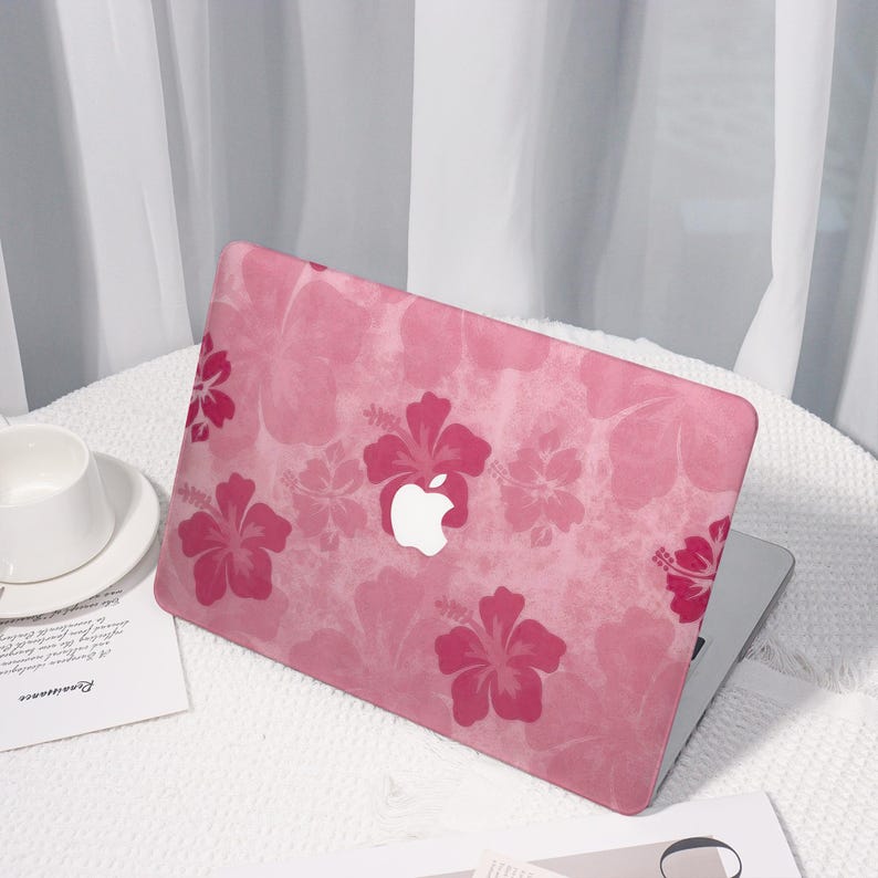 May include: Pink laptop case with a floral hibiscus pattern. The case features a repeating design of pink hibiscus flowers on a lighter pink background. The Apple logo is visible in white. The laptop is on a white surface.