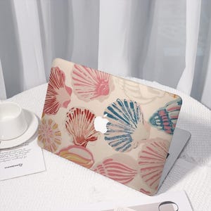 May include: A laptop with a seashell pattern in shades of pink, red, and blue on a beige background. The laptop is open, revealing the Apple logo. The design features various seashell illustrations.