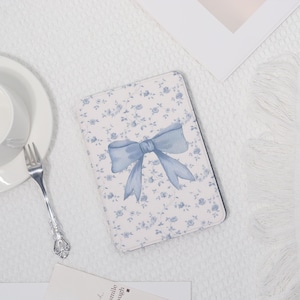 May include: A rectangular notebook with a white background and a blue floral pattern. A large blue bow is centered on the cover. The notebook is surrounded by a white plate, a fork, and a small card.