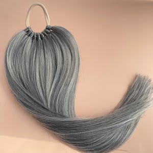 May include: A long, gray hair extension with a rubber band at the top. The hair is tied together in a ponytail style.