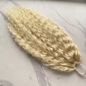 May include: A platinum blonde hair extension piece featuring long, tightly curled strands. The hair is gathered at one end and secured with a small, circular elastic band, creating a ponytail-like structure with a voluminous, wavy texture.