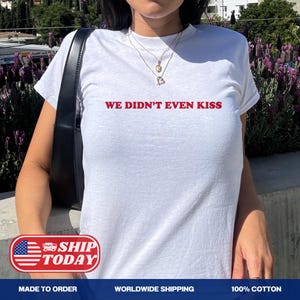 May include: White t-shirt with the red text "WE DIDN'T EVEN KISS". The shirt has a crew neck and short sleeves. The model is wearing a necklace with a heart pendant. The background features purple flowers.