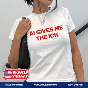 Op de afbeelding: Een wit T-shirt met korte mouwen en de tekst AI GIVES ME THE ICK in dikke rode hoofdletters op de borst. Het shirt wordt gecombineerd met een zwarte schoudertas en een goudkleurige ketting met een hartvormige hanger.