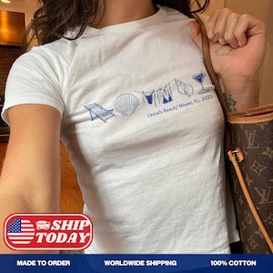 以下が含まれることがあります： 青いビーチをテーマにしたグラフィックが特徴の白いTシャツ。デザインには、ビーチチェア、貝殻、ビキニ、カクテルが含まれています。シャツのテキストは「Chloe's Beach/ Miami, FL, 2025」と書かれています。綿100％。