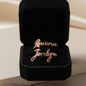 Könnte beinhalten: Ein roségoldener Ring mit den Namen "Rowena" und "Jordyn" in einer schwarzen Samt-Ringbox. Der Ring ist in einer kursiven Schriftart. Die Ringbox ist geöffnet und zeigt den Ring im Inneren. Der Hintergrund ist neutral.