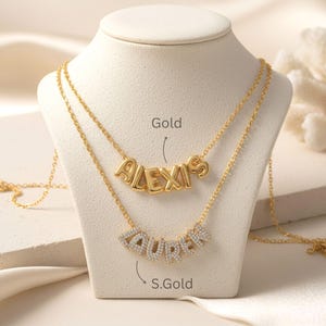 May include: Two gold-toned necklaces displayed on a stand. The top necklace features the name ALEXIS in smooth, rounded gold letters. The bottom necklace features the name LAUREN in letters encrusted with small, clear, sparkling stones.