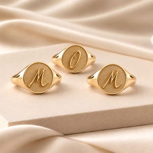 Peut inclure: Trois bagues chevalières en or avec un anneau poli et une face circulaire mate. Deux bagues présentent la lettre M gravée en écriture cursive, tandis que la bague centrale affiche la lettre O gravée dans le même style d'écriture.