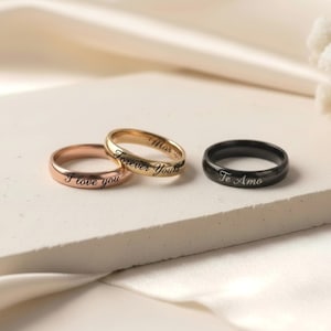 Peut inclure: Trois bagues en m&eacute;tal avec des anneaux lisses. Une bague en or rose est grav&eacute;e avec le texte I love you. Une bague en or est grav&eacute;e avec le texte Forever Yours. Une bague noire est grav&eacute;e avec le texte Te Amo en &eacute;criture cursive.