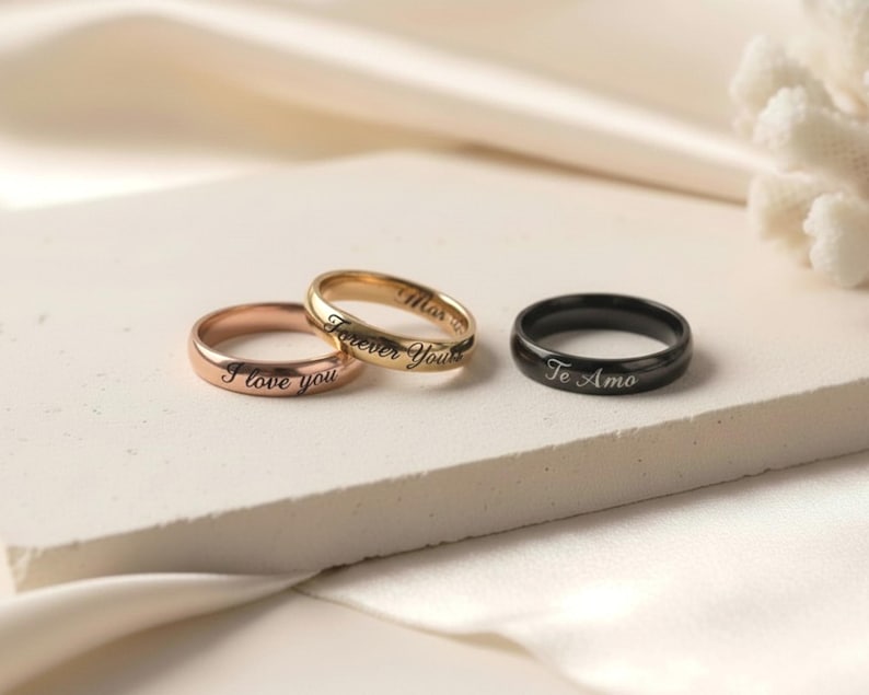 Peut inclure: Trois bagues en m&eacute;tal avec des anneaux lisses. Une bague en or rose est grav&eacute;e avec le texte I love you. Une bague en or est grav&eacute;e avec le texte Forever Yours. Une bague noire est grav&eacute;e avec le texte Te Amo en &eacute;criture cursive.