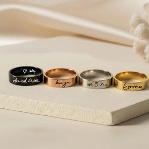 Peut inclure: Quatre bagues en m&eacute;tal aux tons noir, or rose, argent et or. Chaque bague pr&eacute;sente une inscription grav&eacute;e personnalis&eacute;e. La bague noire porte l'inscription "All our love" avec un c&oelig;ur et "Haley", l'or rose "love you", l'argent "we love you" et l'or "Emma".