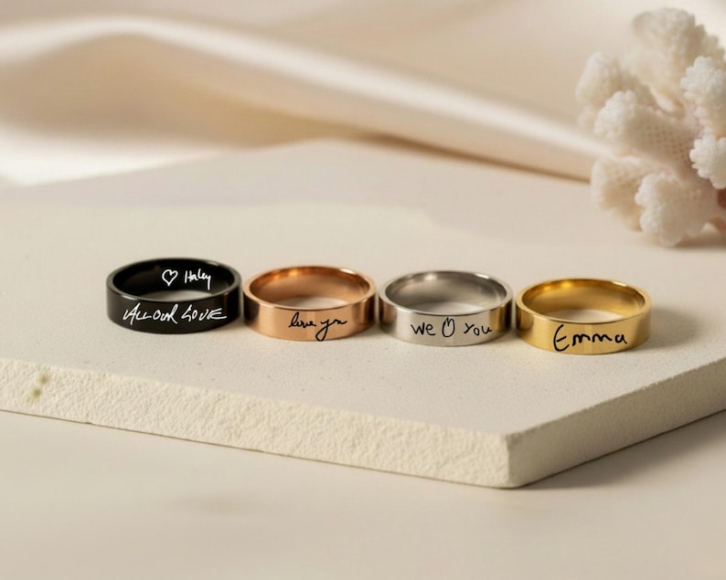Peut inclure: Quatre bagues en m&eacute;tal aux tons noir, or rose, argent et or. Chaque bague pr&eacute;sente une inscription grav&eacute;e personnalis&eacute;e. La bague noire porte l'inscription "All our love" avec un c&oelig;ur et "Haley", l'or rose "love you", l'argent "we love you" et l'or "Emma".