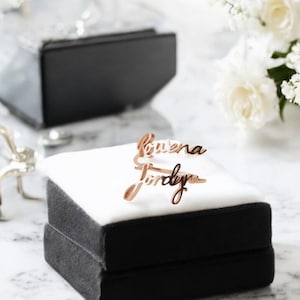 Könnte beinhalten: Zwei Ringe aus Roségold mit den eingravierten Namen "Luvena" und "Jordyn". Die Ringe werden in einer weißen Samtbox präsentiert.