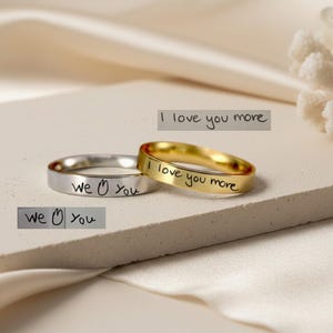 Peut inclure: Deux bagues en m&eacute;tal, une argent&eacute;e et une dor&eacute;e, chacune pr&eacute;sentant une gravure manuscrite. La bague argent&eacute;e porte l'inscription "we [symbole c&oelig;ur] you" et la bague dor&eacute;e porte l'inscription "I love you more."