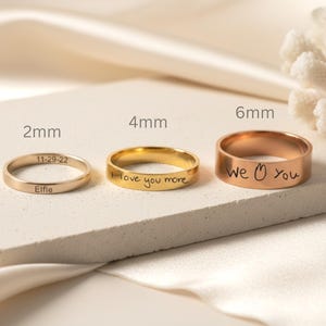 Könnte beinhalten: Drei Metallringe mit unterschiedlichen Breiten von 2mm, 4mm und 6mm. Der 2mm Ring ist mit 11-29-22 und Elfie graviert. Der 4mm Ring trägt den Text I love you more. Der 6mm Ring zeigt den Text we [Herzsymbol] you.