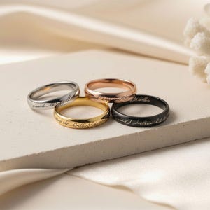 May include: Four rings in silver, rose gold, gold, and black, each with engraved script. The rings are displayed on a neutral surface with a soft, blurred background. The rings are a simple band design.