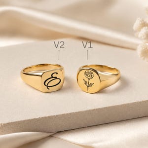 May include: Two gold-toned signet rings. The ring labelled V2 features a black engraved cursive letter E on a square face. The ring labelled V1 features a black engraved floral design on a round face. Both rings have a polished metallic finish.